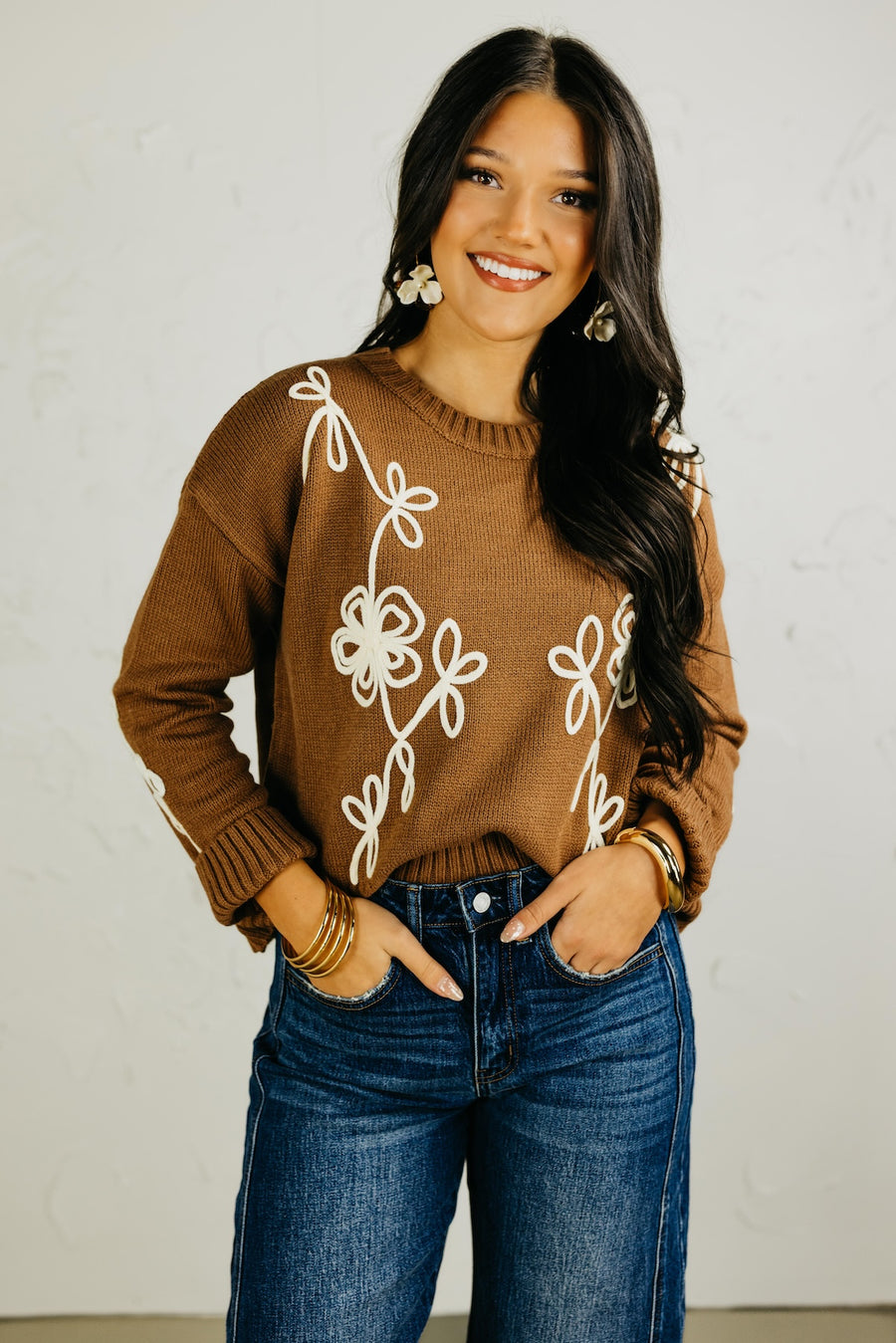 Woman wearing a brown sweater with white floral patterns and blue jeans against a plain background