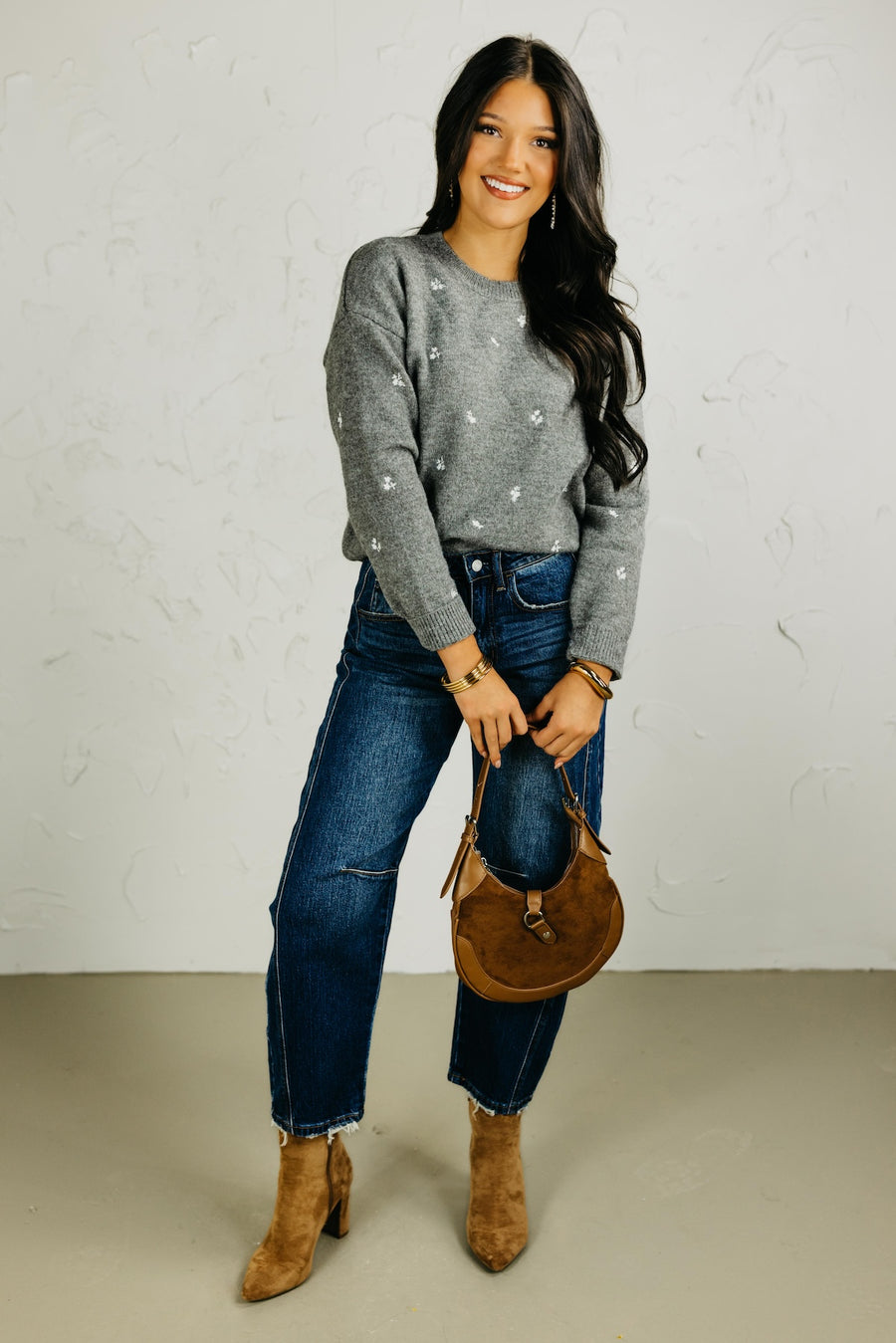 Woman wearing a gray sweater with floral patterns, blue jeans, and holding a brown bag against a plain background.
