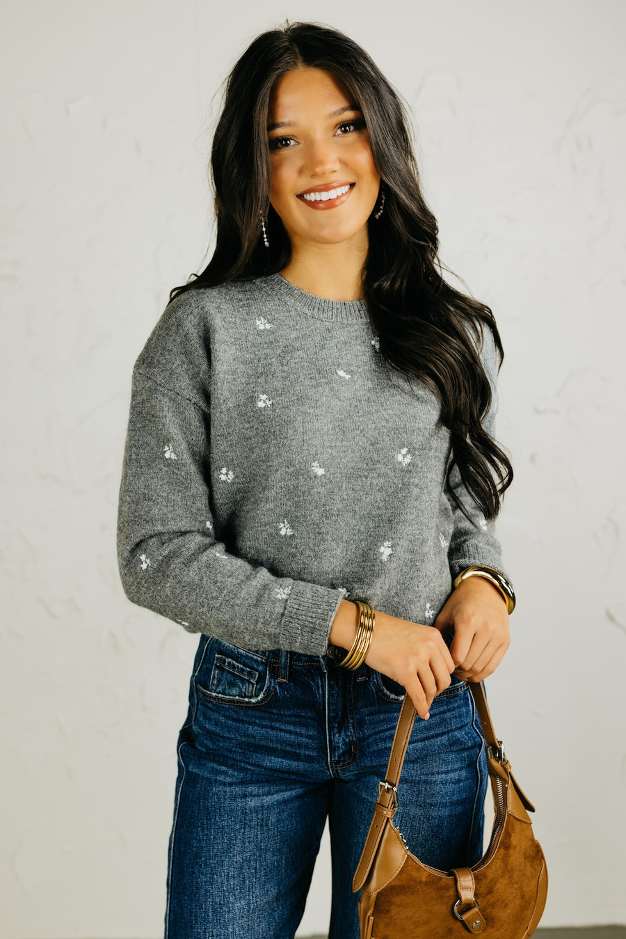 Woman wearing a gray sweater with floral patterns, blue jeans, and holding a brown bag against a plain background.