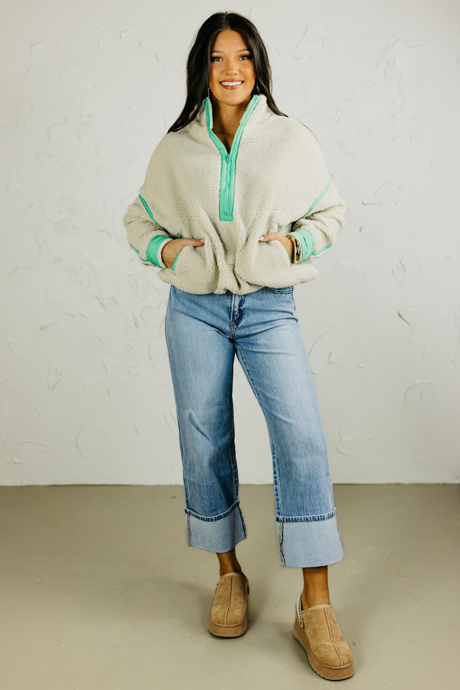 Woman wearing a beige fleece jacket with green trim, blue jeans, and brown shoes against a white background.